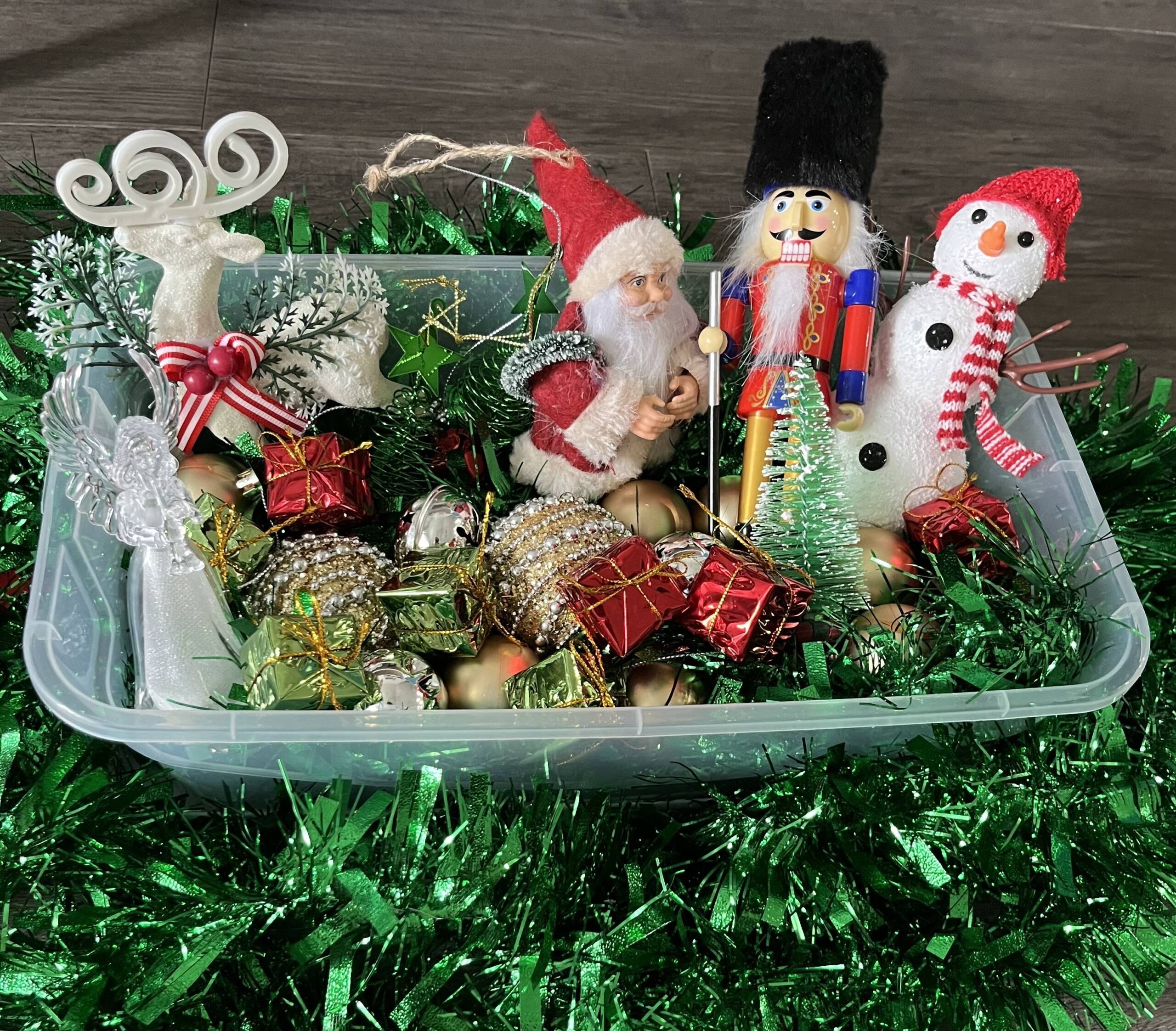Sensory box that includes items for the Christmas season. Minature snowman, nutcracker, Santa, plastic lightup angel, tiny presents, a reindeer.