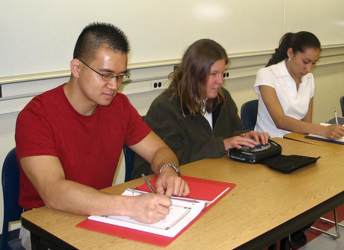 Anna sits between a male student and a female student. All 3 are taking notes - Anna with her portable note taker and the others with pen and paper.