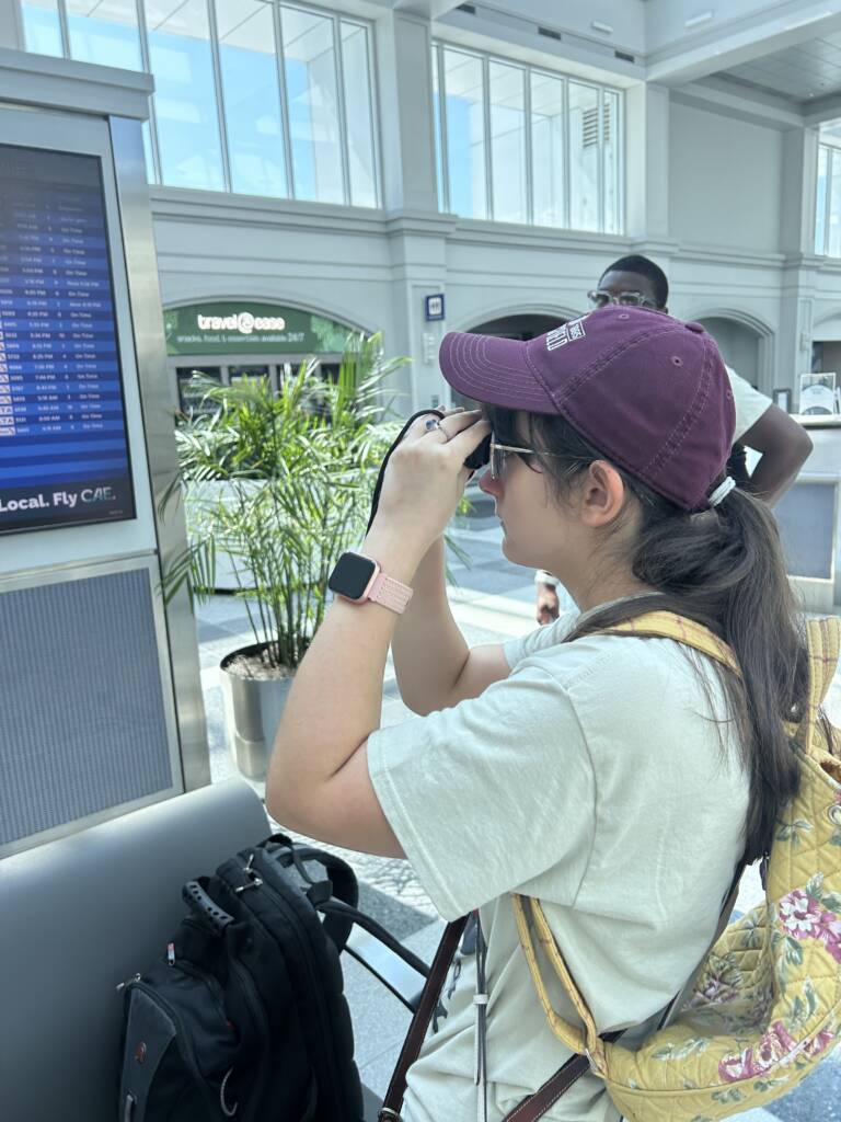 A female traveler uses a monocular to locate the gate for her flight on an airport monitor. 