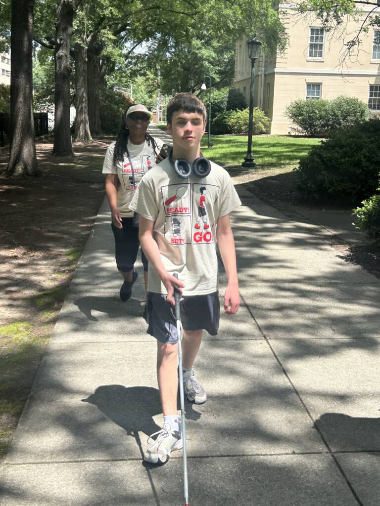 A male traveler walks down a sidewalk using a white cane. A woman follows behind him. Both are wearing program t-shirts. 