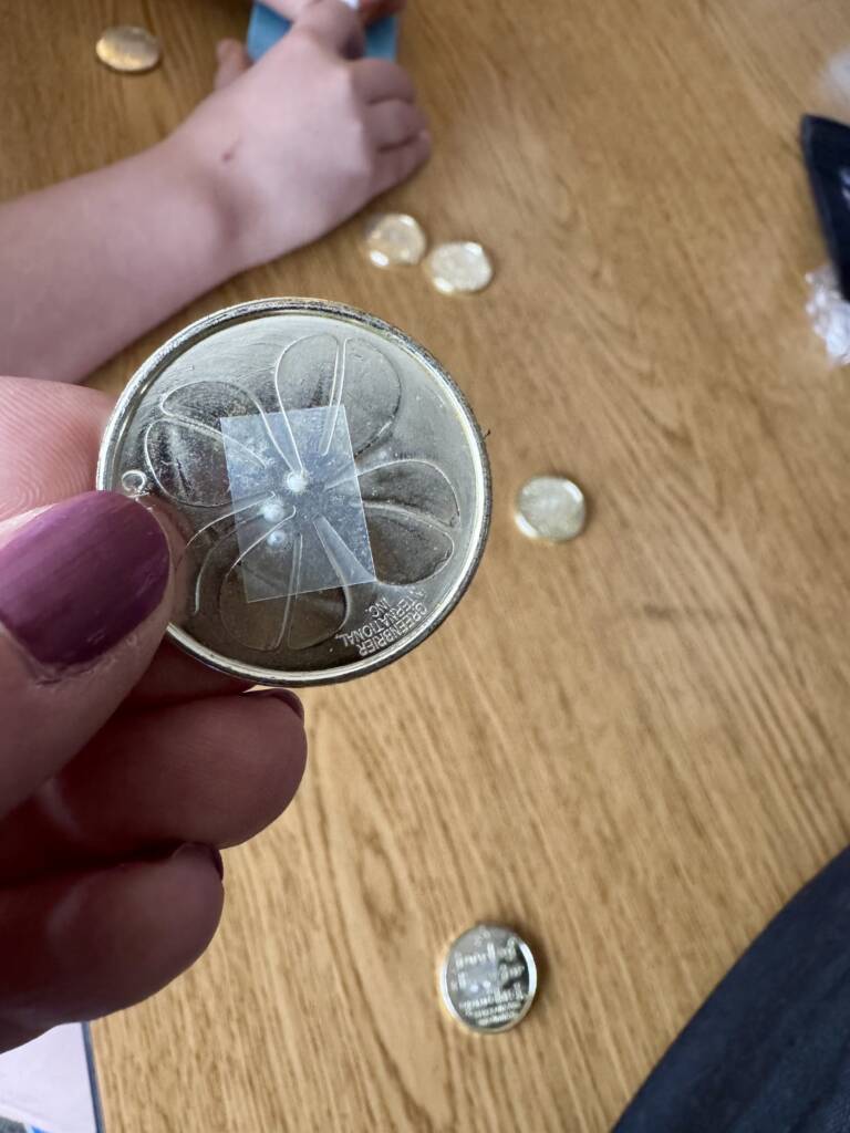 Gold coin that has a four leaf clover imprint and a braille sticker with the letter "s". 