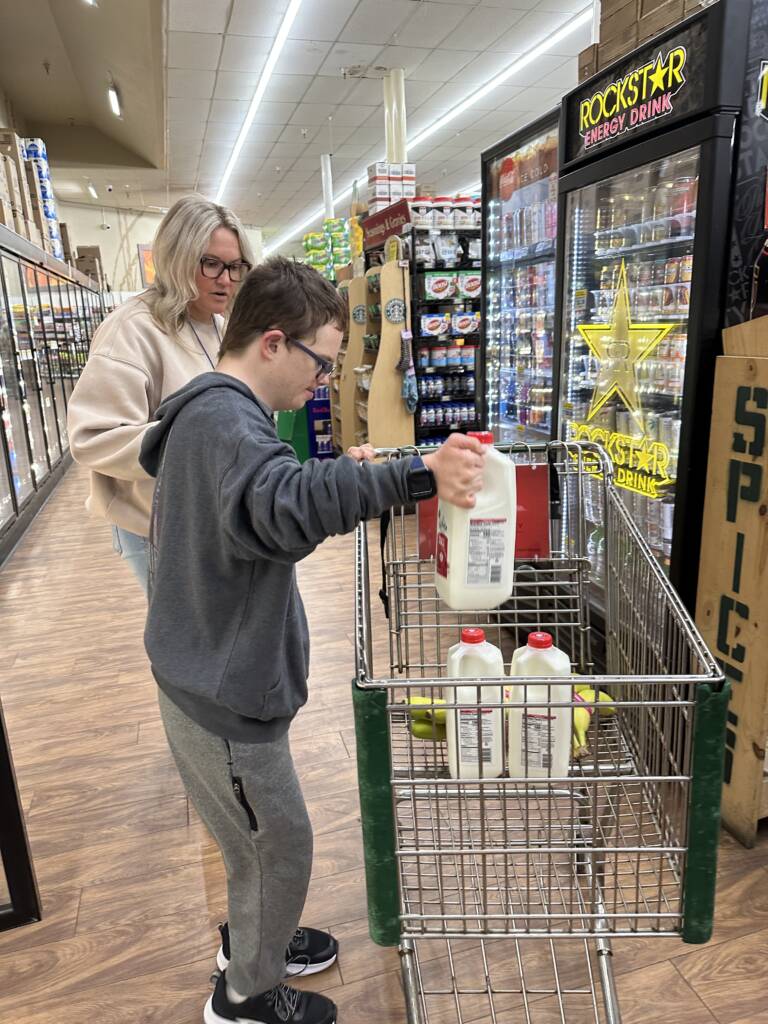 Student is putting a 1/2 gallon of milk in the shopping cart with the teacher observing. 