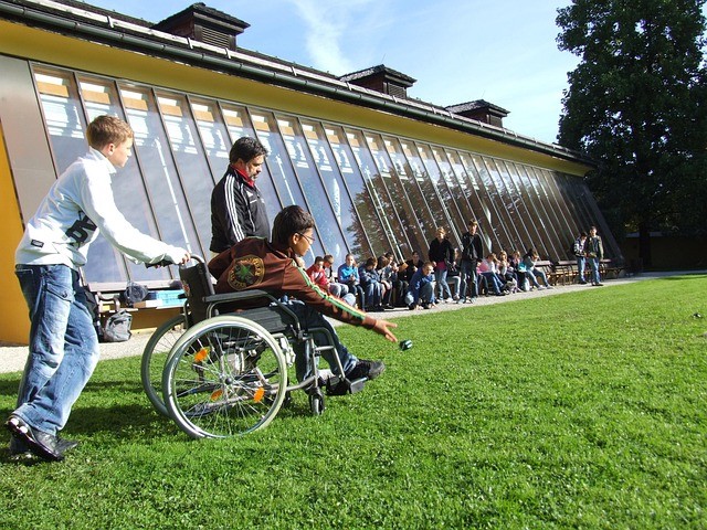 A group of students outside on a grassy lawn. One student is pushing a student in a wheel chair while the student rolls a ball.