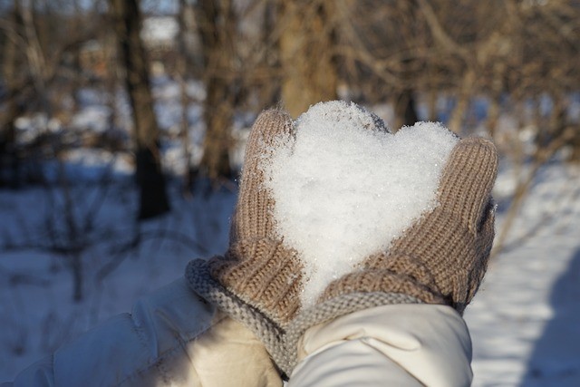 Hands with mittens on holding a heart shaped snowball outside.