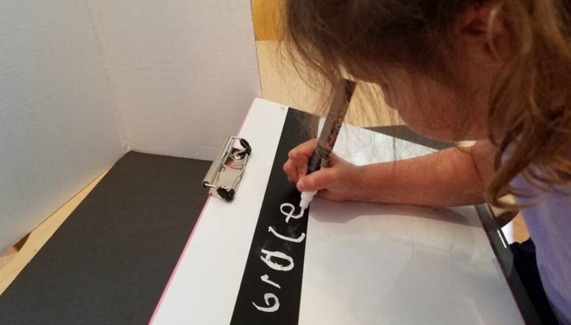 Student writing with a white marker on a black strip of paper on a white slant board.