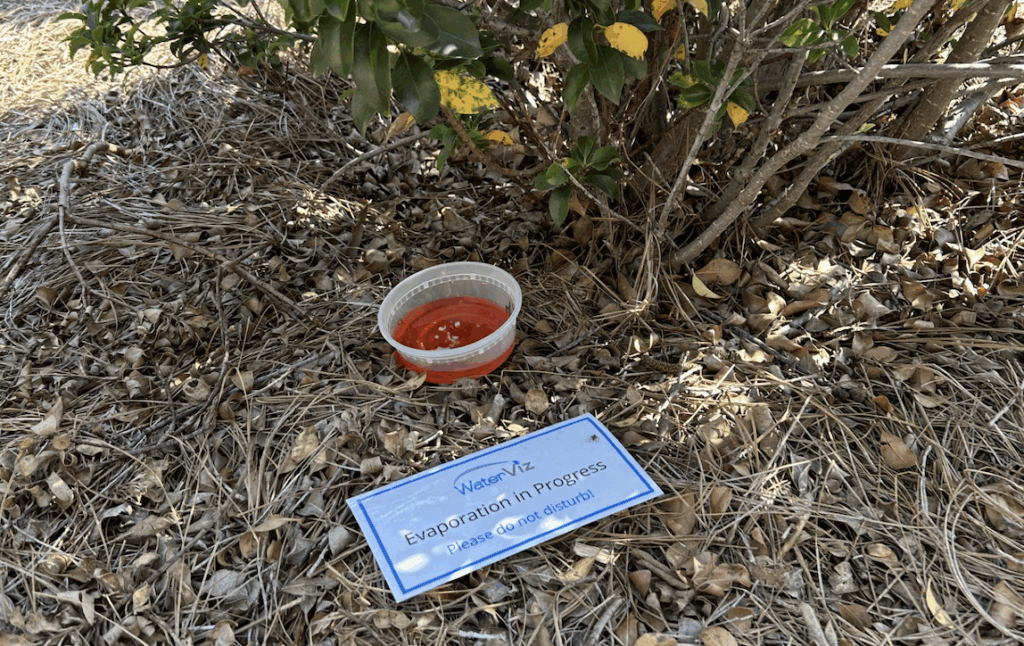 Photo of an evaporation dish containing colored water placed on the ground in the shade under a bush. Beside the dish is a sign that reads “Evaporation in Progress Please Do Not Disturb!)